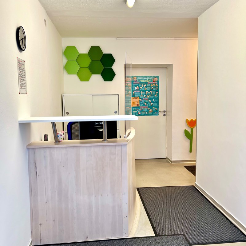 Empfangsbereich der Praxis: helle Holz-Anmeldung mit grünen Wabenpaneelen an der Wand